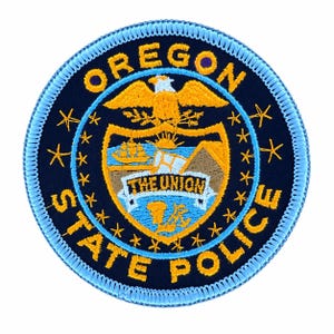 May include: A circular embroidered patch with a dark blue background and a light blue stitched border. It features the Oregon state seal in yellow and blue thread, including an eagle, a ship, and the text OREGON STATE POLICE and THE UNION.