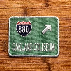 May include: Green and white embroidered patch with a white arrow pointing right. The patch has a blue and white interstate highway sign with the number 880 and the text "Interstate" above the number. The text "Oakland Coliseum" is below the sign.