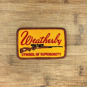 May include: A yellow rectangular patch featuring the word "Weatherby" in red script above a brown rifle with a scope. The text "SYMBOL OF SUPERIORITY" is below the rifle, within a brown border.