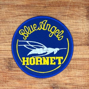 May include: A circular patch with a blue background and yellow embroidered text that reads "Blue Angels" and "Hornet". A white and blue stylized image of an aircraft is in the center. The patch is on a wooden surface.