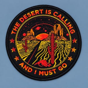 Round black desert patch with orange “The Desert Is Calling and I Must Go” lettering, featuring a sunset, cacti, desert road, stars, and skull design on a blue-gray background.