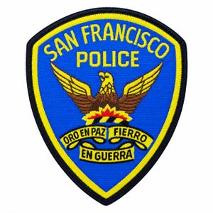 May include: A shield-shaped embroidered patch with a blue background and yellow border. It features the text SAN FRANCISCO POLICE in yellow, a central eagle graphic in brown and yellow, and a yellow ribbon with the text ORO EN PAZ FIERRO EN GUERRA.