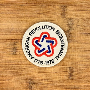 Puede incluir: Un parche circular con el texto "AMERICAN REVOLUTION BICENTENNIAL 1776-1976" en blanco. El parche presenta un diseño de estrella roja y azul en el centro, con un borde blanco. El parche está sobre una superficie de madera.