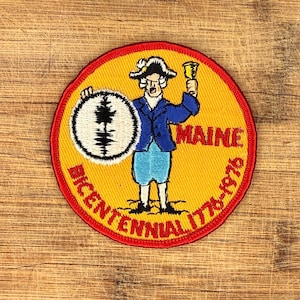 Puede incluir: Un parche redondo amarillo con un borde rojo que presenta una figura colonial sosteniendo una campana y un escudo con un diseño de árbol. También están presentes los textos "MAINE" y "BICENTENNIAL 1776-1976".