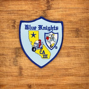 May include: A shield-shaped embroidered patch with a light blue background and dark blue border. It features the text Blue Knights, a yellow map of California with a star, a motorcycle, and a smaller shield emblem with a knight figure.