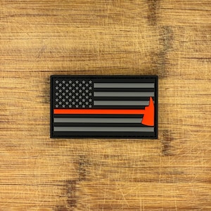 May include: A rectangular patch with a design of the American flag. The flag is black and grey with a red line and a red axe. The stars are white. The patch is on a wooden surface.