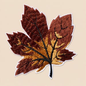 May include: An embroidered iron-on patch shaped like a maple leaf. The leaf features a color gradient transitioning from deep brown at the outer edges to a warm golden yellow toward the center, with prominent black stitched veins throughout.