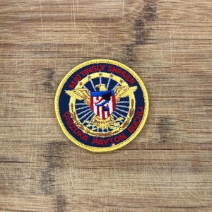 May include: A circular embroidered patch with a dark blue background and a yellow border. The center features a golden eagle with wings spread, holding a shield with red and white stripes. The text MATTINGLY SHRIVER ONIZUKA PAYTON BUCHLI is stitched in red.