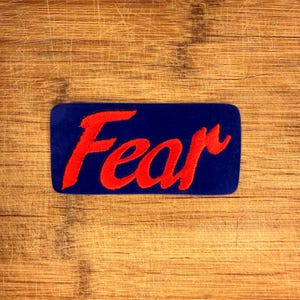 Könnte beinhalten: Ein rechteckiger blauer Aufnäher mit dem Wort "Fear" in roter, kursiver Schrift gestickt. Der Aufnäher hat abgerundete Ecken und ist auf einem Holzgrund mit sichtbarer Maserung und Kratzern angebracht.