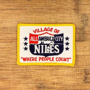 May include: Embroidered patch with a white background, yellow border, and text that reads "VILLAGE OF NILES ALL AMERICA CITY WHERE PEOPLE COUNT." The patch features red and navy blue text and three stars on each side.