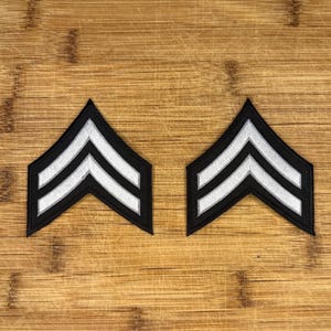 以下が含まれることがあります： 木目調の背景に、2つの黒と白の刺繍されたシェブロン（おそらく軍の階級章）が表示されています。各シェブロンは、黒い背景に2本の白いストライプがあり、古典的な上向きのシェブロンの形に配置されています。