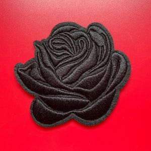May include: Black embroidered rose patch with detailed stitching. The patch is in the shape of a rose and is set against a bright red background. The patch is likely for clothing or accessories.