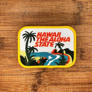 May include: Embroidered patch with a yellow border and a white background. The patch features palm trees, ocean waves, and the text "HAWAII THE ALOHA STATE" in red. The patch is rectangular with rounded corners.