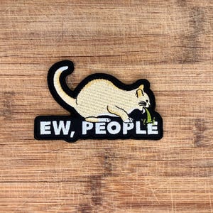 Puede incluir: Parche bordado con un gato de color beige claro con contorno negro, vomitando verde. El gato está posicionado sobre un rectángulo negro con las palabras "EW, PEOPLE" en letras mayúsculas blancas. Sobre una superficie de madera.