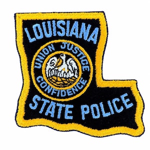 Puede incluir: Un parche bordado con la forma del estado de Luisiana con fondo negro y borde amarillo. Presenta el texto LOUISIANA y STATE POLICE en azul claro, con un emblema circular central que muestra un pelícano blanco y el texto UNION JUSTICE CONFIDENCE.