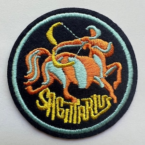 May include: A round, black felt patch with a blue border. The center features an embroidered image of the Sagittarius zodiac sign in orange, yellow, and blue. The word "Sagittarius" is embroidered in yellow below the image.