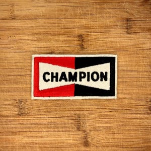 May include: A rectangular patch featuring the word "CHAMPION" in black, bold lettering. The patch is split into three sections: a white central area, a red section on the left, and a black section on the right. It rests on a wooden surface.