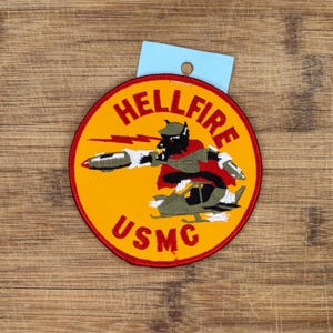 Op de afbeelding: Een ronde, geborduurde patch met de tekst "HELLFIRE" en "USMC" in het rood op een gele achtergrond. De patch toont een cartoonfiguur met een raket en een helikopter. Het heeft een rode rand en een blauw label.