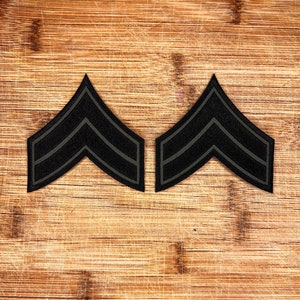 May include: Two black fabric military rank insignia patches with two chevron stripes on a wooden surface.