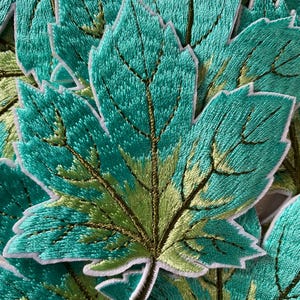 May include: A close-up of several teal and green embroidered fabric maple leaves. The leaves have a white outline and are arranged in a pile.