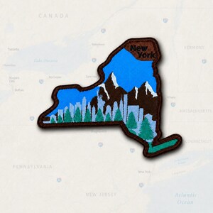 May include: A fabric patch shaped like the state of New York with a dark brown border. The design features a bright blue sky, dark brown mountains, a light blue city skyline with the Statue of Liberty, and green pine trees. The text NEW YORK is printed.