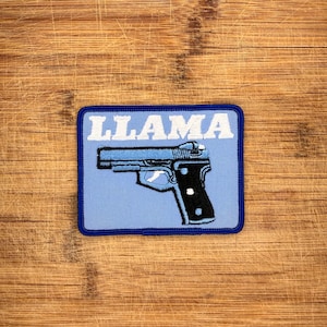 Puede incluir: Un parche bordado rectangular con fondo azul claro y borde azul oscuro. La palabra LLAMA está bordada en letras mayúsculas blancas sobre un gráfico de una pistola con corredera azul claro y empuñadura negra con puntos blancos.
