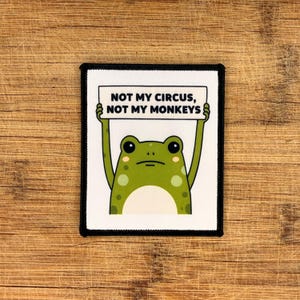 May include: Square patch featuring a green frog with a sign saying "NOT MY CIRCUS, NOT MY MONKEYS." The frog has large eyes and spots. The patch has a black border and is on a wooden surface.