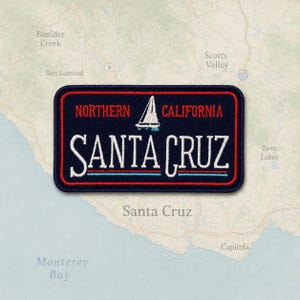 May include: A rectangular embroidered patch with a dark blue background and a red border. It features the white text "NORTHERN CALIFORNIA" above a white sailboat icon, with the large white text "SANTA CRUZ" and thin red and blue lines below.