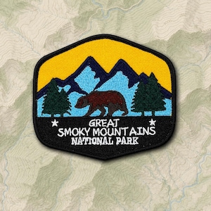 May include: A hexagonal embroidered patch featuring a brown bear walking in front of light blue mountains and dark green pine trees against a yellow sky. The black bottom section displays the white text GREAT SMOKY MOUNTAINS NATIONAL PARK.