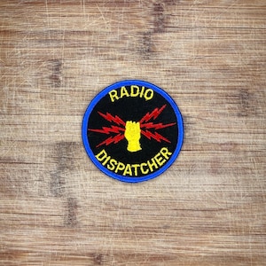 Pode incluir: Um emblema bordado circular com fundo preto e borda azul. O centro apresenta um gráfico de um punho amarelo rodeado por raios vermelhos. O texto RADIO DISPATCHER está bordado em letras maiúsculas amarelas ao redor do centro.