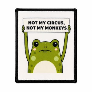 May include: A rectangular embroidered patch with a white background and a thick black border. The design features a green frog with light green spots holding a white sign above its head that reads, NOT MY CIRCUS, NOT MY MONKEYS, in black text.