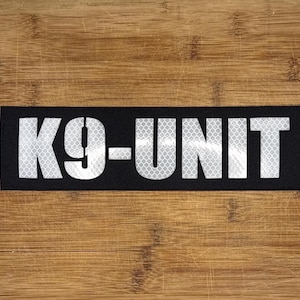 May include: A black rectangular patch with the text "K9-UNIT" in reflective white letters. The patch is set against a wooden background.