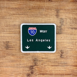 May include: A rectangular patch resembling a green highway sign. The sign reads "Interstate 10 West Los Angeles" with white text and arrows. The patch has a white border and a blue Interstate 10 logo.
