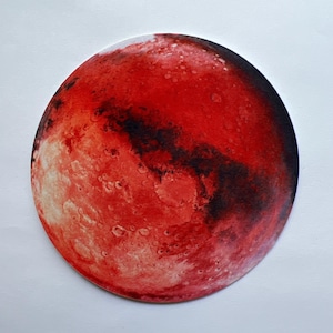 May include: A round, red and black illustration of the planet Mars. The image features a detailed depiction of the planet's surface, with craters and dark patches. The artwork is set against a white background.