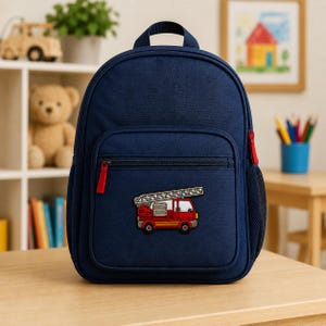 Red fire truck embroidered kids patch displayed on the front pocket of a navy blue child’s backpack in a bright playroom setting, with toys, books, and art supplies softly blurred in the background.