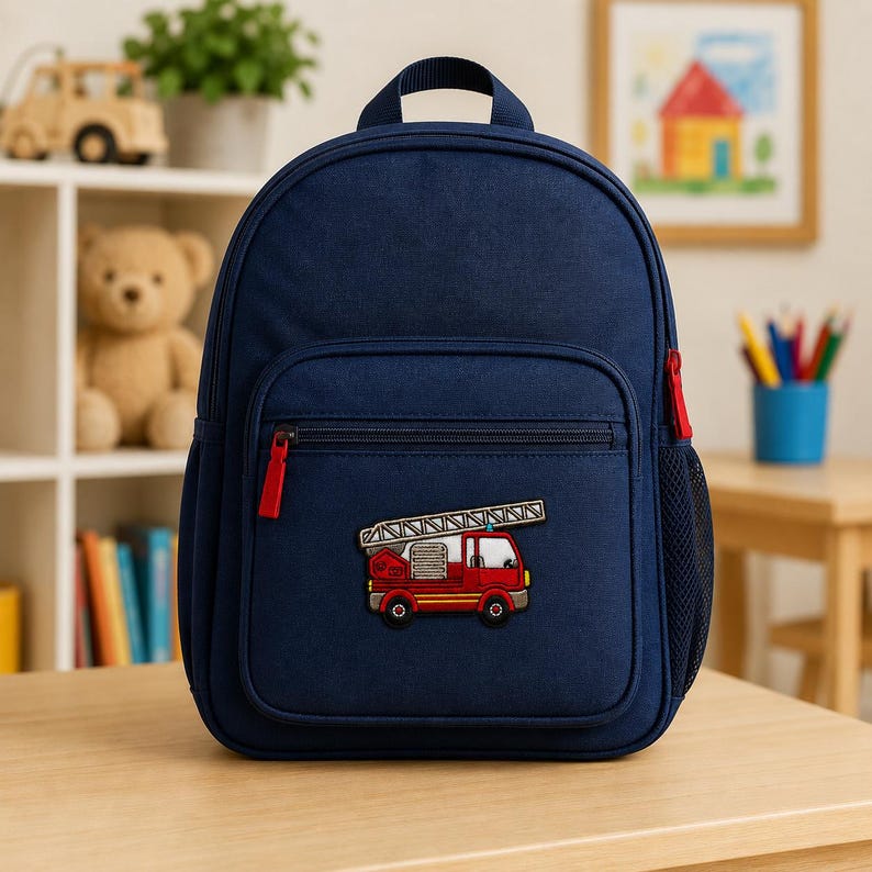 Red fire truck embroidered kids patch displayed on the front pocket of a navy blue child’s backpack in a bright playroom setting, with toys, books, and art supplies softly blurred in the background.