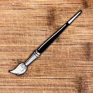 May include: A black and white embroidered patch of a paintbrush on a wooden background.