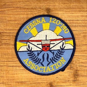 Puede incluir: Parche redondo azul con el texto amarillo "CESSNA 120-140 ASSOCIATION". El parche presenta un diseño estilizado de avión con ala roja, nube blanca y sol.