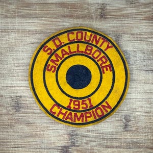 Op de afbeelding: Een vintage gele vilten patch met de tekst "S.D. COUNTY SMALLBORE 1951 CHAMPION" in rode letters. De patch heeft een cirkelvormig ontwerp met concentrische ringen en een donkerblauw midden. De patch ligt op een houten oppervlak.