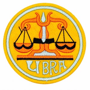 Op de afbeelding: Een ronde geborduurde patch met een gele achtergrond en een witte rand. In het midden staat een oranje weegschaal met zwarte accenten. Onder de weegschaal staat het woord LIBRA geborduurd in witte letters met een zwarte omlijning.