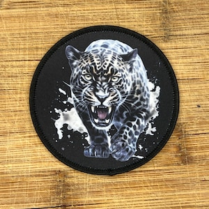 May include: A black and white jaguar with a fierce expression, printed on a round black patch. The jaguar is walking towards the viewer with its mouth open, showing its teeth.