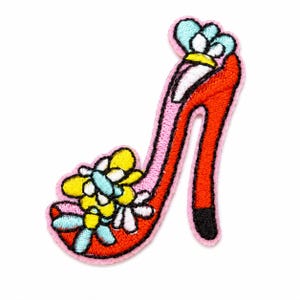 May include: An embroidered patch in the shape of a red high-heeled shoe with a light pink border. The shoe is decorated with yellow and light blue flower-shaped accents on the toe and a light blue flower detail at the top of the heel.