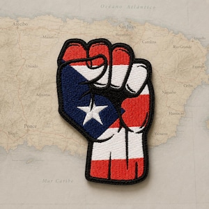 May include: An embroidered patch in the shape of a raised fist, featuring the red, white, and blue colours of the Puerto Rican flag. The design includes a blue triangle with a single white star on the thumb side and horizontal stripes.