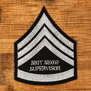 May include: Black and white embroidered patch with the text "SHIT SHOW SUPERVISOR". The patch has a chevron design with three white stripes on a black background, resembling military rank insignia.
