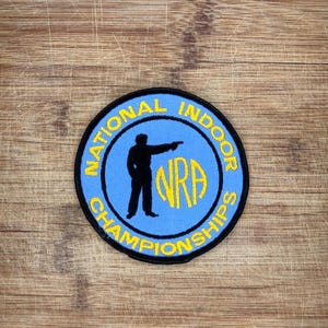 May include: A circular embroidered patch with a light blue background and a black border. It features a black silhouette of a person aiming a firearm, the yellow text NRA, and the yellow text NATIONAL INDOOR CHAMPIONSHIPS encircling the center.