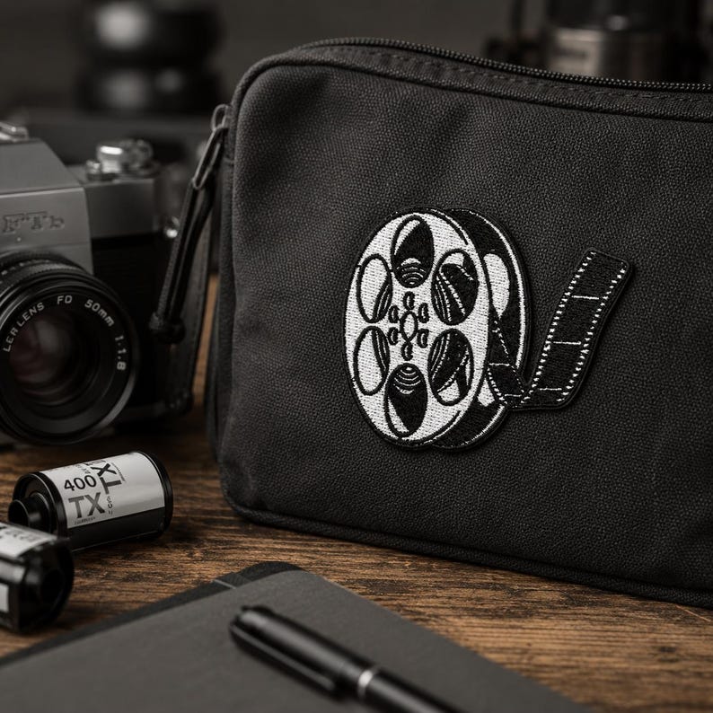 Vintage film reel embroidered patch attached to a black camera bag with film rolls and photography gear nearby.