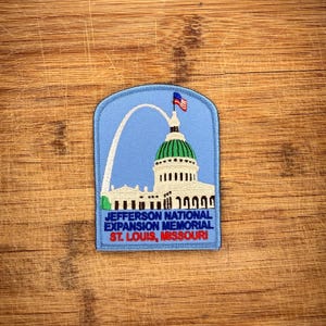 Pode incluir: Um emblema bordado em azul claro apresentando o Gateway Arch de St. Louis e o edifício Old Courthouse com uma pequena bandeira americana no topo. O texto diz JEFFERSON NATIONAL EXPANSION MEMORIAL ST. LOUIS, MISSOURI em azul e vermelho.