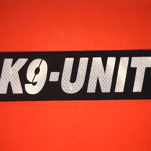 May include: Black rectangular patch with the text "K9-UNIT" in reflective white lettering. The patch is set against a bright orange background. The letters have a textured, grid-like pattern.