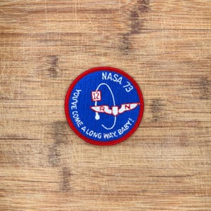 May include: A circular embroidered patch with a blue background and a red border. The design features a white stylized wing graphic, the number 12 in a red square, and the text NASA 73 YOU'VE COME A LONG WAY BABY! in white lettering.