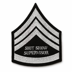 May include: A black embroidered patch shaped like a military chevron with three white stripes and the text SHIT SHOW SUPERVISOR stitched in white thread. The patch features a pointed top and a curved bottom edge with a thick black border.